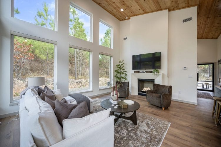 Living room windows on two walls looking onto pines, sofa and fireplace at the far end