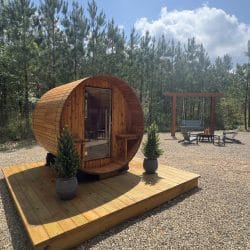 Cedar barrel sauna on a wood platform with a porch swing frame and fire pit chairs behind it
