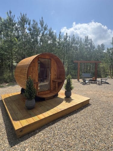 Cedar barrel sauna on a wood platform with a porch swing frame and fire pit chairs behind it
