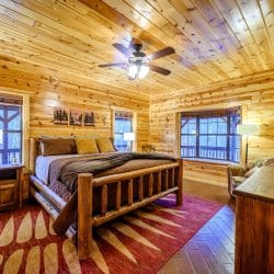 Bed with a log frame and red rug, ceiling fan and a bay of windows facing the deck