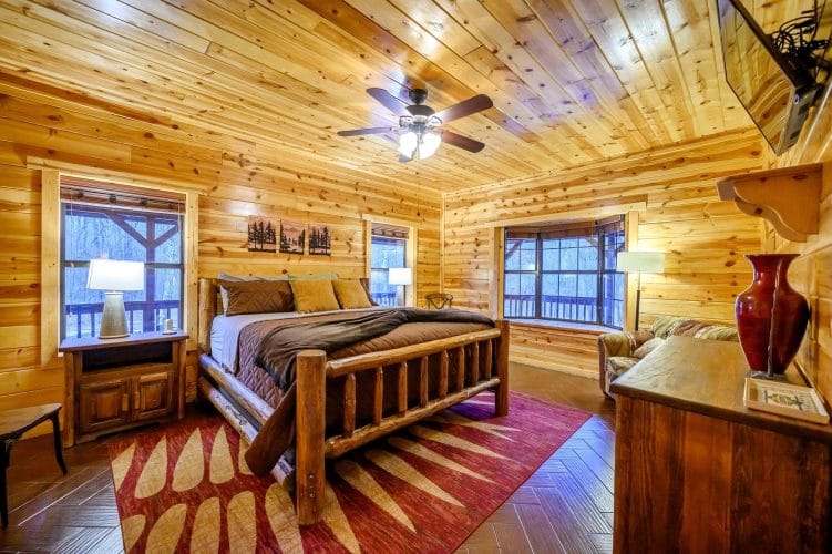Bed with a log frame and red rug, ceiling fan and a bay of windows facing the deck