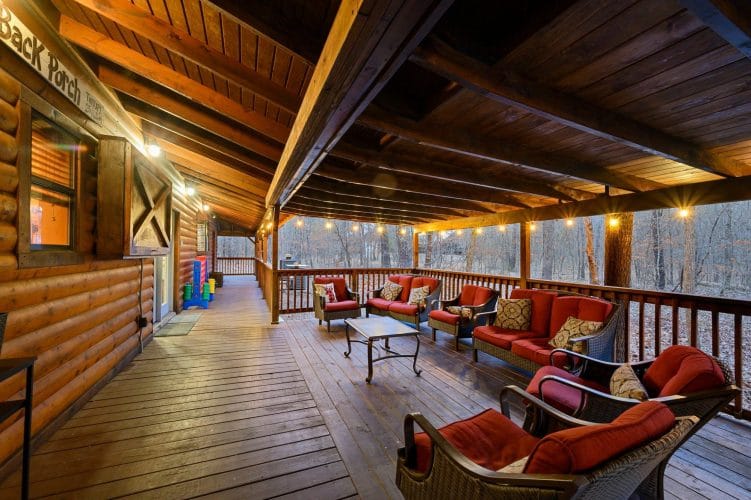 Covered deck with a sofa and chairs with red cushions around a coffee table, string lights overhead