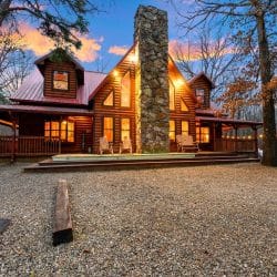 Front of the log cabin with a stone chimney, red roof and lit windows at dusk