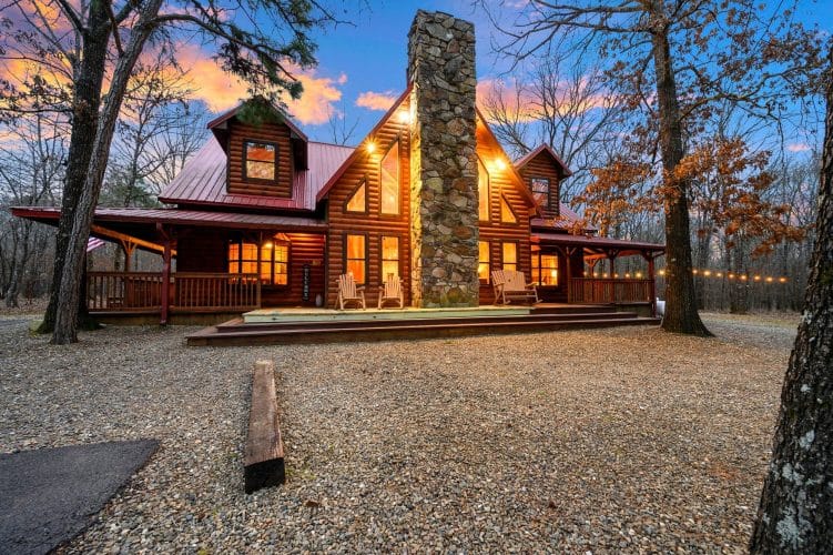 Front of the log cabin with a stone chimney, red roof and lit windows at dusk