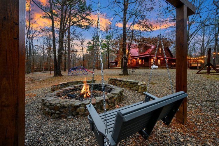 Fire pit with a bench swing in the foreground and the cabin behind it