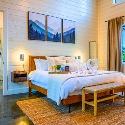 King bed with white bedding and a towel decoration on it, mountain art above and a wood bench at the foot
