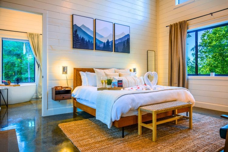 King bed with white bedding and a towel decoration on it, mountain art above and a wood bench at the foot