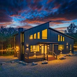Front of the dark-sided modern cabin with a sloped roof and lit windows at sunset, with string lights along the edge of the lot