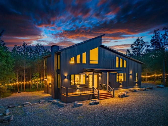 Front of the dark-sided modern cabin with a sloped roof and lit windows at sunset, with string lights along the edge of the lot