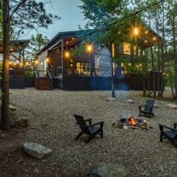Fire pit with Adirondack chairs on gravel and the lit cabin behind