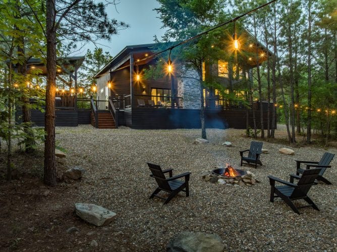 Fire pit with Adirondack chairs on gravel and the lit cabin behind