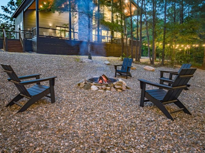Fire pit with black Adirondack chairs on gravel and the lit deck behind