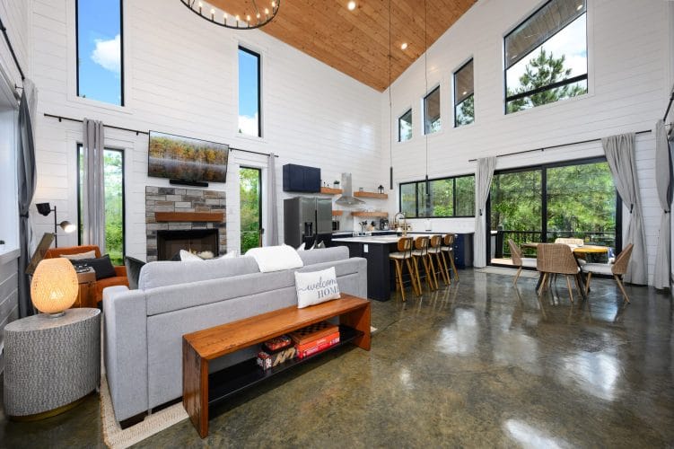Open living area under a vaulted ceiling with a gray sofa and wood bench, the kitchen and dining table behind