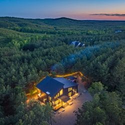 Aerial of the cabin and gravel drive in the forest at dusk, string lights along the lot