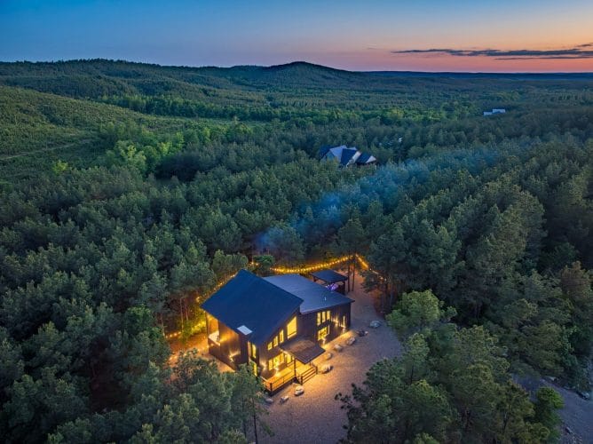 Aerial of the cabin and gravel drive in the forest at dusk, string lights along the lot